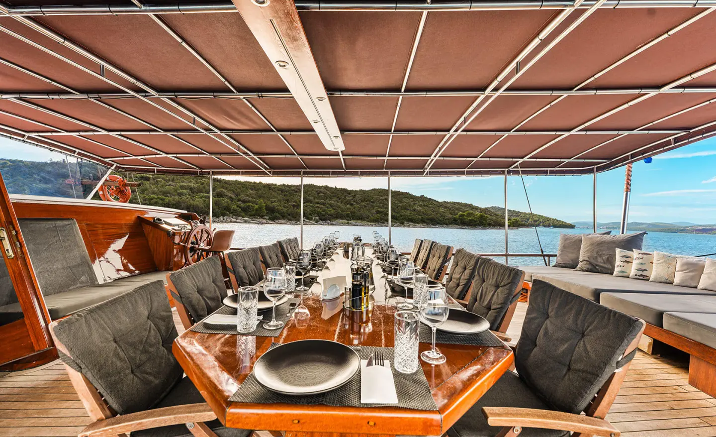 Outdoor yacht dining area with a long table set for a meal under a shaded deck, sunset water view