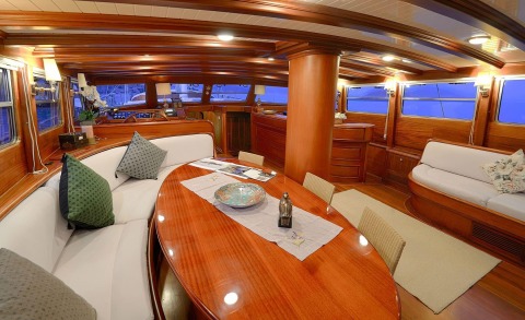 Interior of a wooden yacht with a large oval table, white cushioned seating, and surrounding chairs; helm area in the back and side windows showing night sea