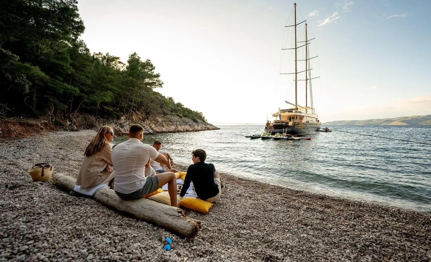 Dalmatino yacht at sunset anchored near rocky shore with passengers on pebble beach