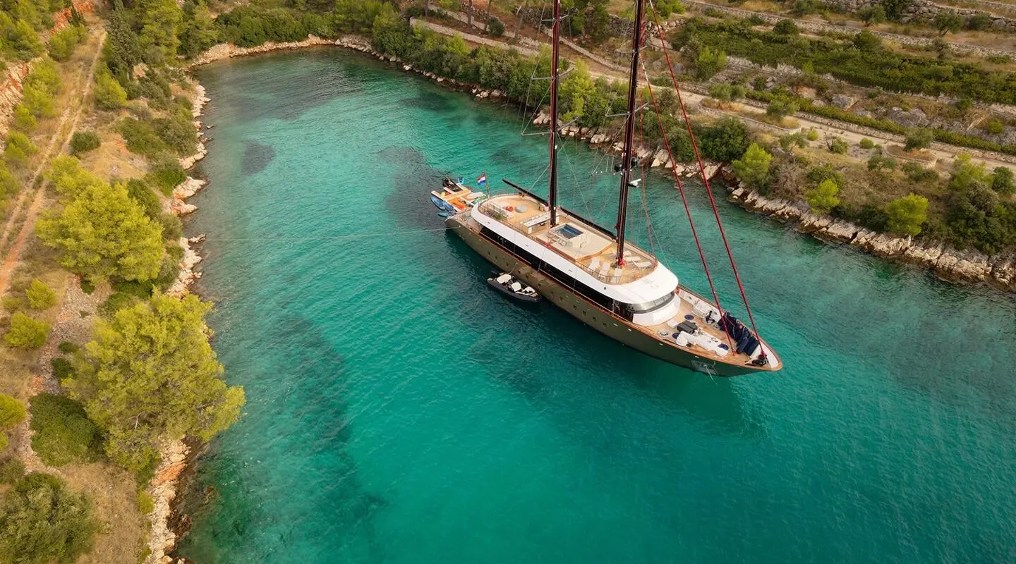 A sleek sailing yacht named Anetta anchored in clear turquoise water beside a stone quay