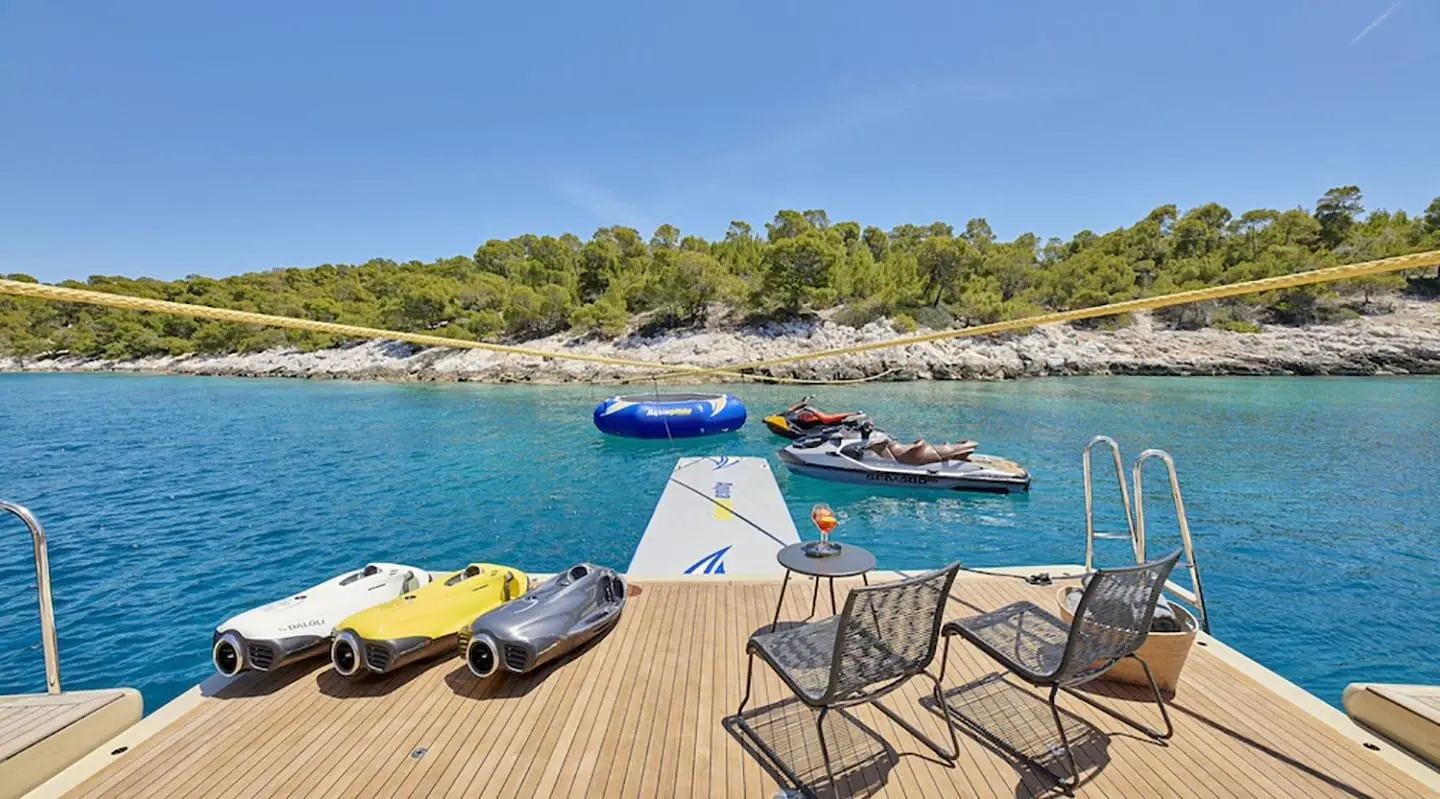 Sunlit yacht deck with lounge chairs, a blue wakeboard boat, and calm turquoise water