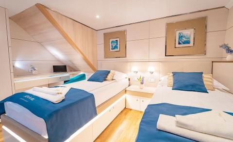 Twin bed cabin with light wood panels, bedside lamps, blue pillows, and a small desk area in a yacht interior