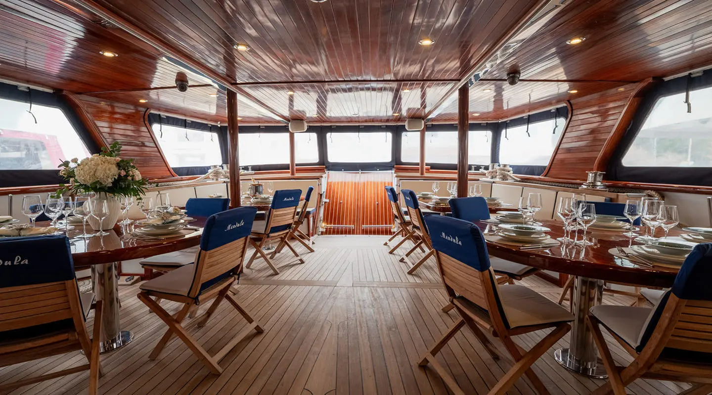 Mendula yacht interior with wooden deck, blue cushions, and seating under canopy at sunset
