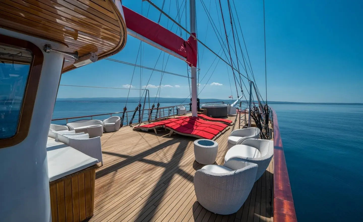 Deck view of Barbara yacht with red sail, wooden planks, and seating cushions