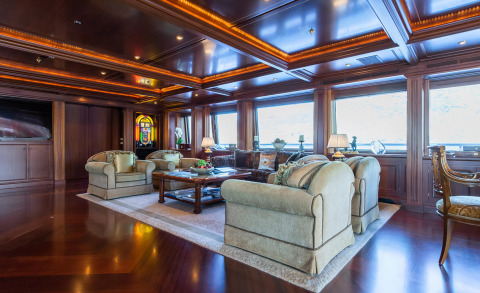 Luxurious 62.5m yacht Apogee interior: plush beige sofas, polished wood panels, warm ambient lighting, large windows, coffee table, and a classic lounge area