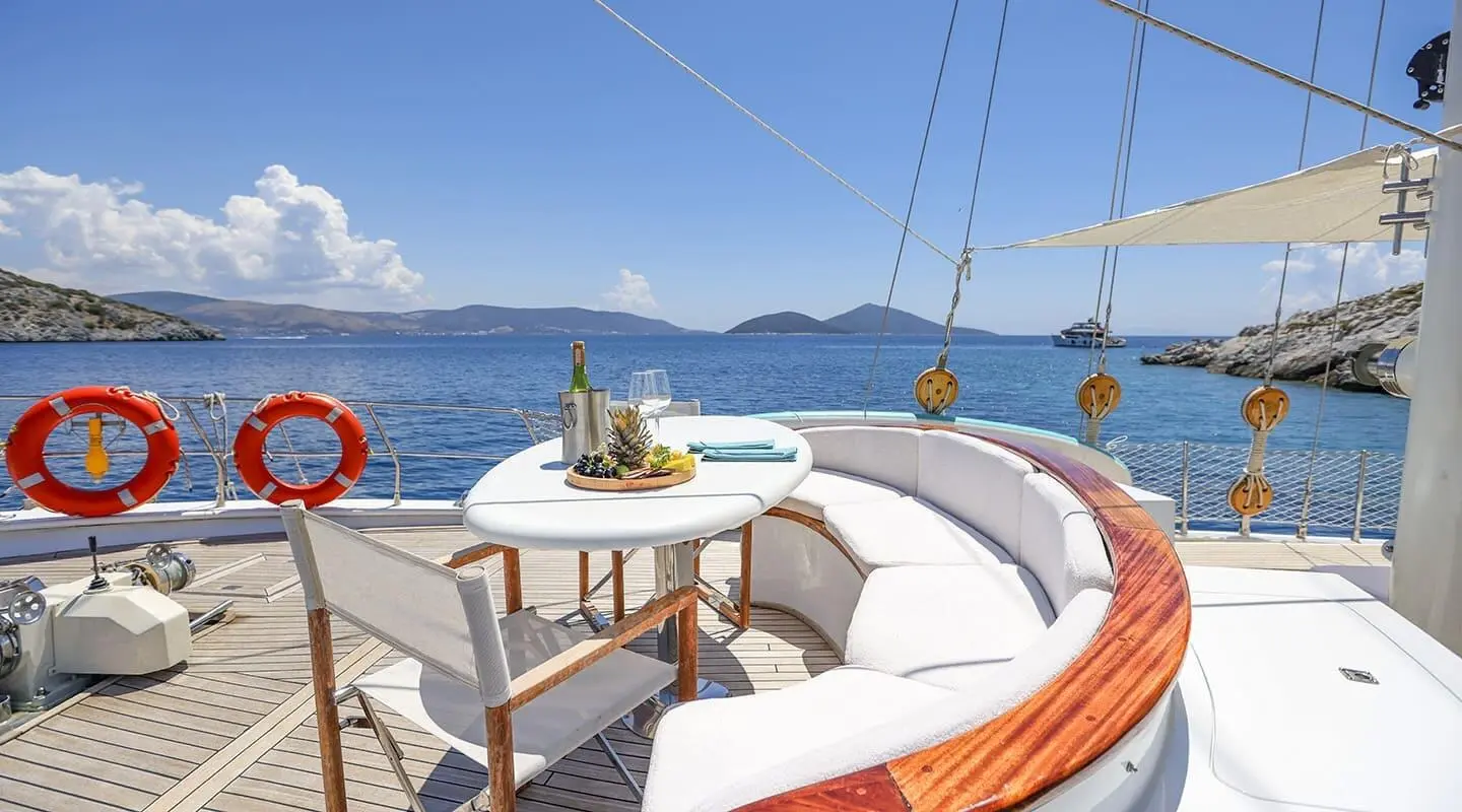 Gazella yacht deck seating area with table, sunlit water, sails in distance