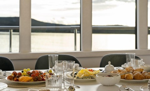 Breakfast spread with croissants on a dining table inside a yacht salon, large windows with a view