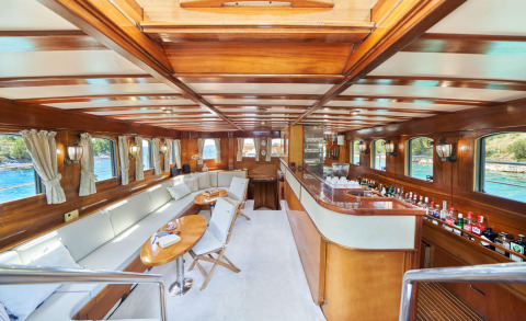 Wood-paneled yacht salon with long white sofa, round wooden tables, bar area on the right and windows along the sides