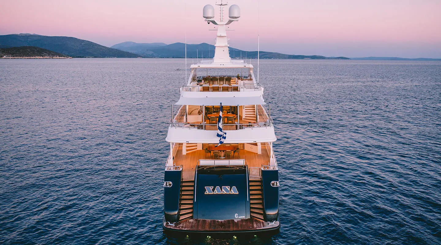 Xana yacht anchored on calm blue water, rear view from above at sunset