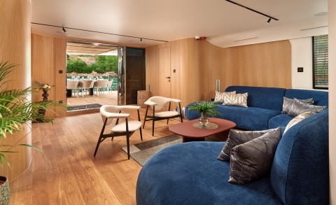 Anetta interior: spacious saloon with blue sectional sofa, cream chairs, wooden walls and deck, open aft doors to a dining area by the water