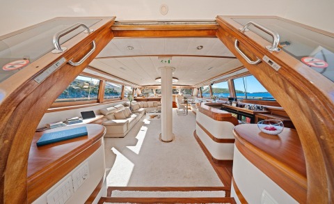 Alessandro I interior: luxurious saloon with cream upholstery and wood trim, panoramic windows, central support column, and teak accents onboard