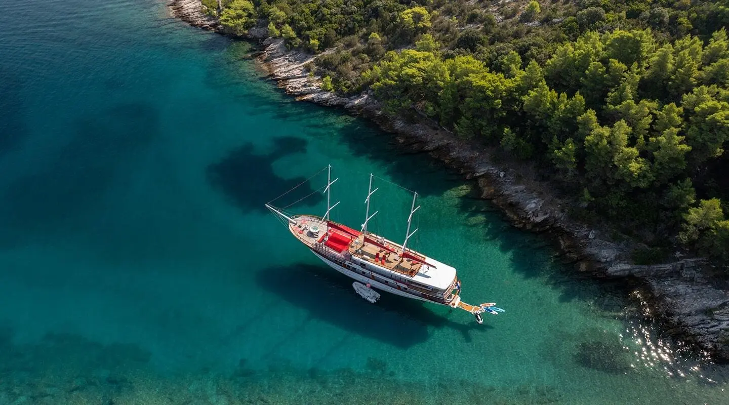 A red-and-white sailing yacht anchored in turquoise water near rocky shoreline