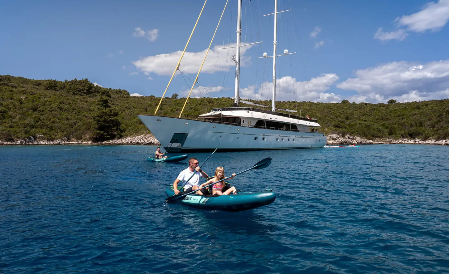 Small inflatable boat with two people approaching a luxurious sailboat anchored near a rocky coastline
