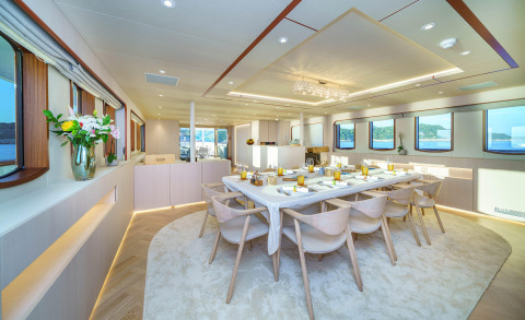 Maia yacht saloon interior with central table and chairs, large windows and wooden furnishings