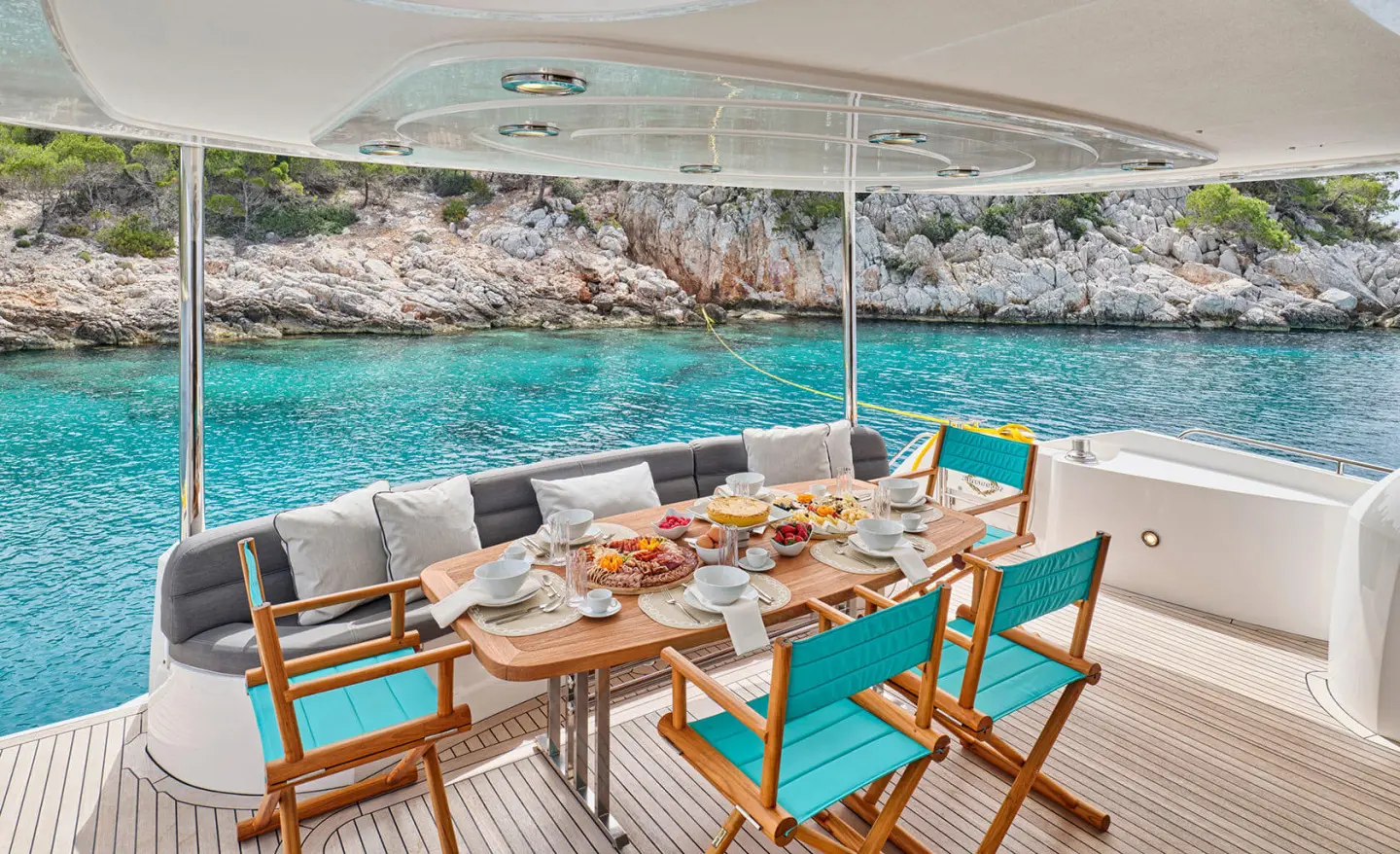 Dining table on a luxury yacht deck with teal chairs, sea and rocky shoreline