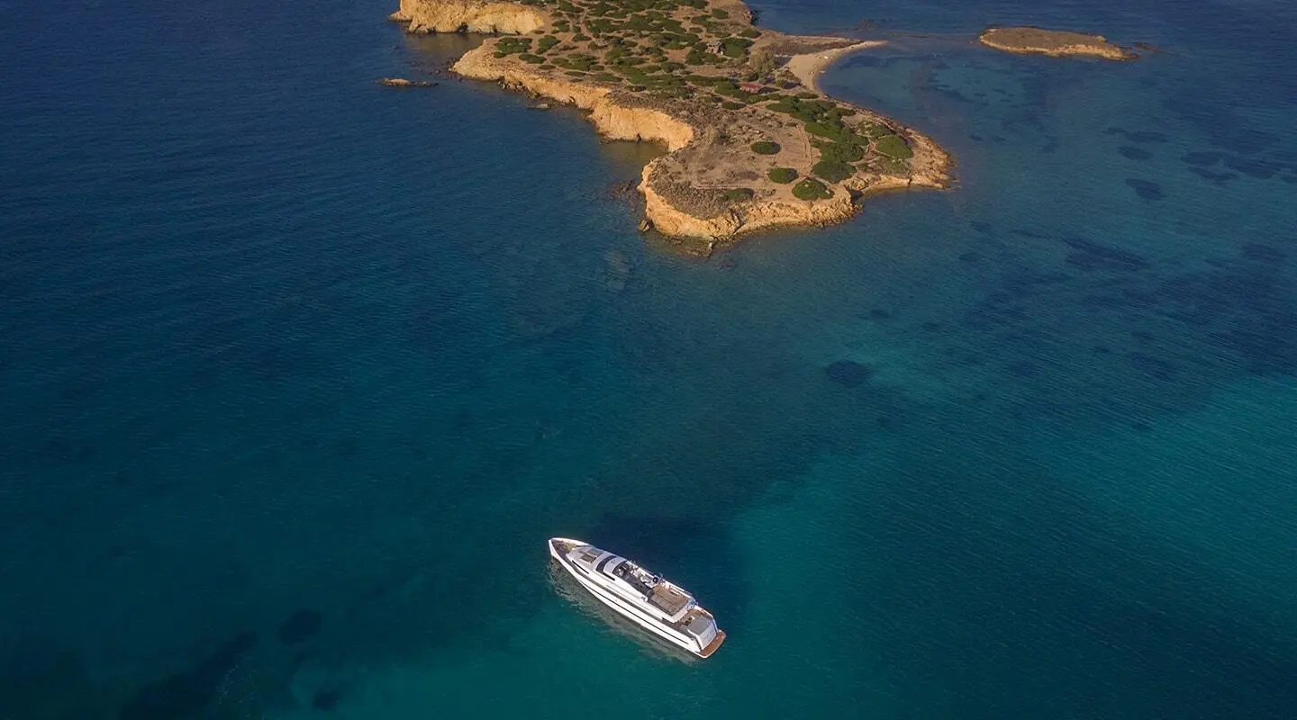 A sleek 34m luxury yacht cruises near a rugged, sunlit coastline island