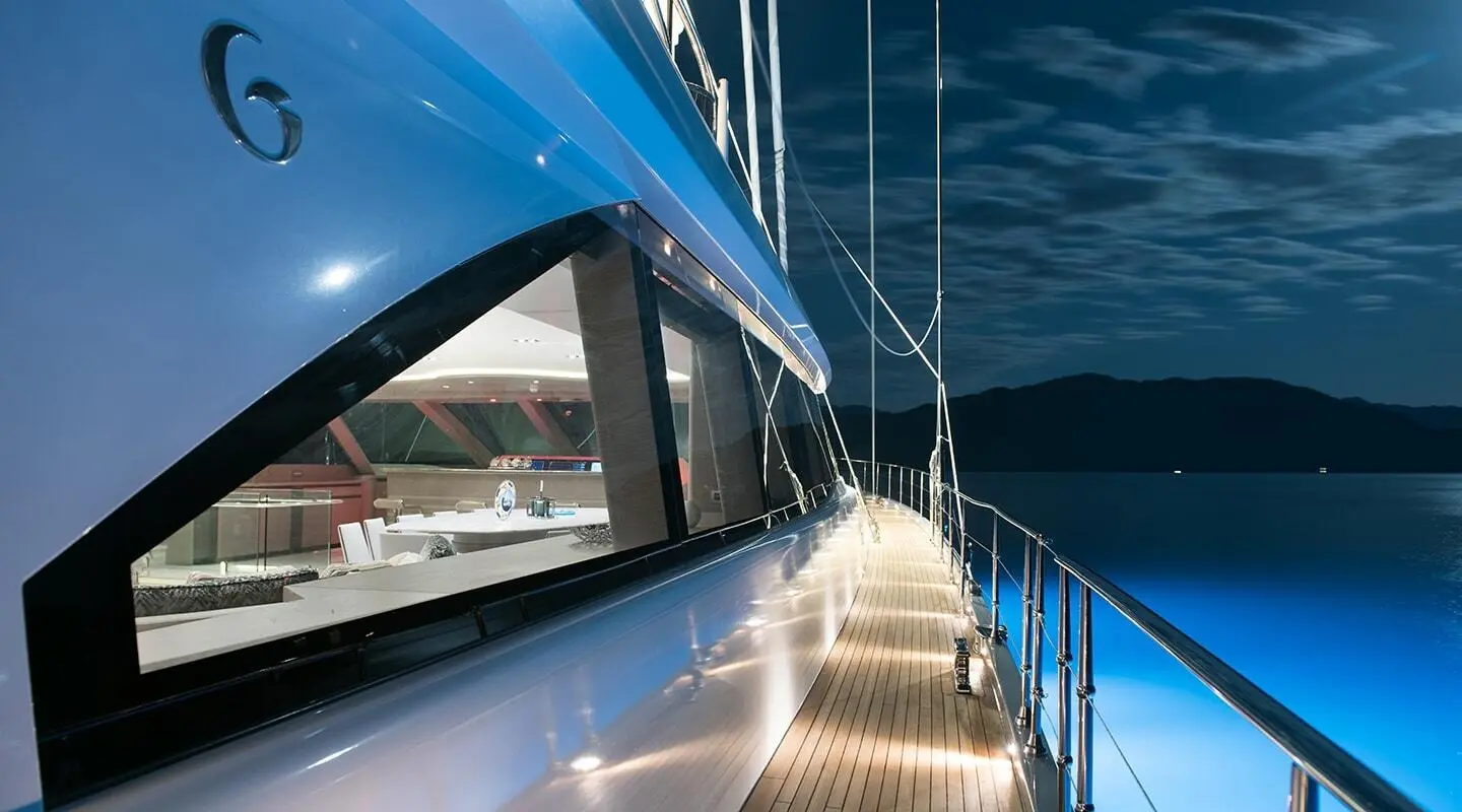 Side deck of a blue yacht with large windows showing an interior dining area