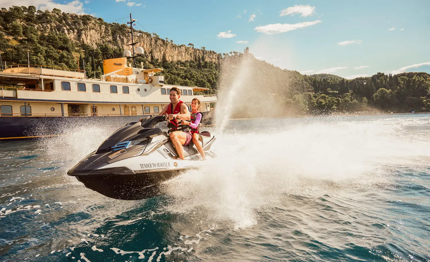 Seagull II jet-skiing near a luxurious yacht anchored by a cliffside coastline