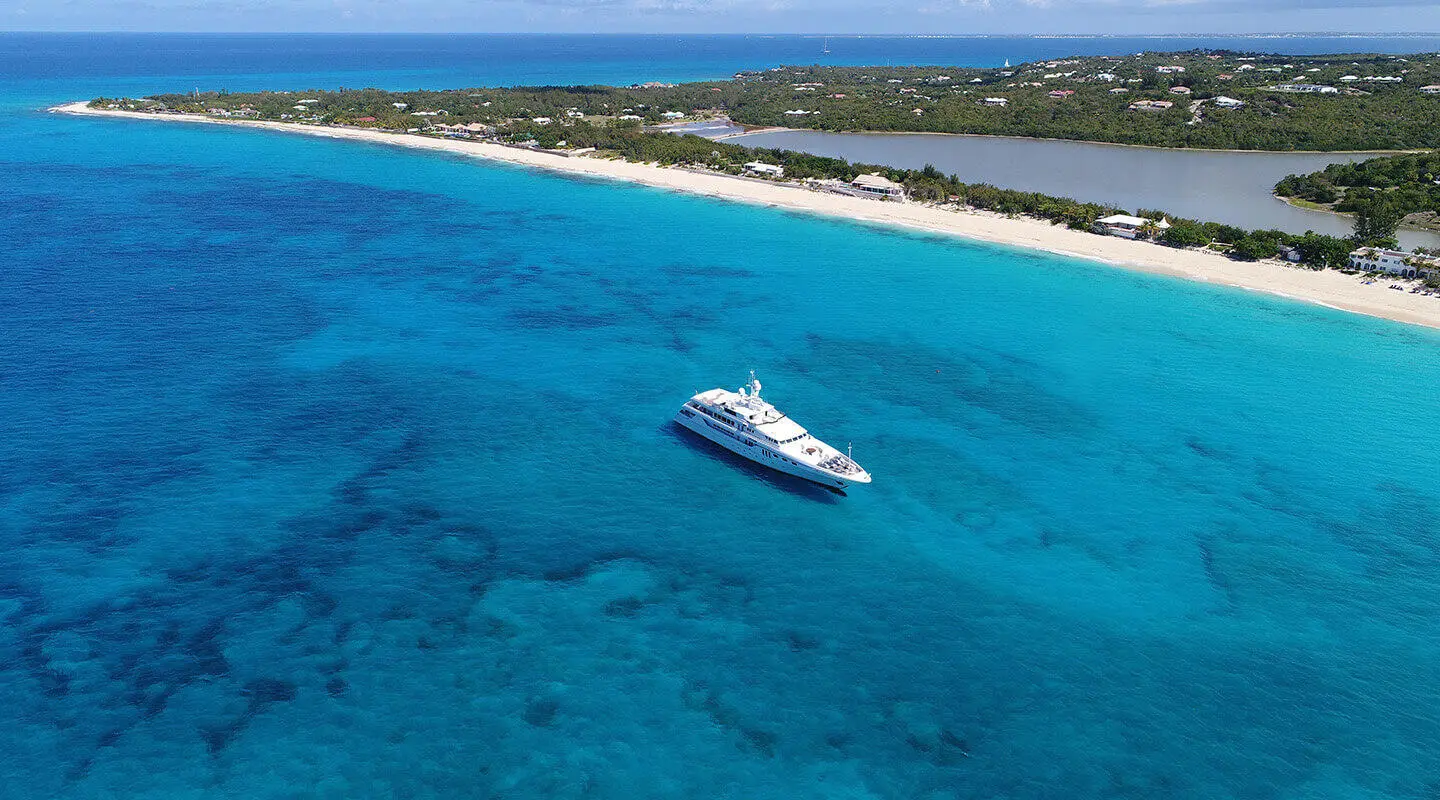 A sleek white yacht named Apogee docks near a sandy coastline in turquoise water
