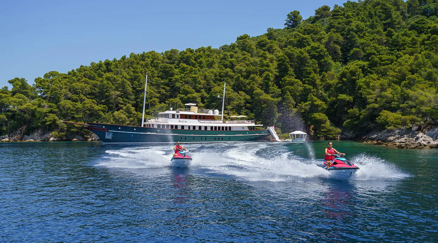 Yacht Maia gliding on calm water near a forested shoreline, wake trailing