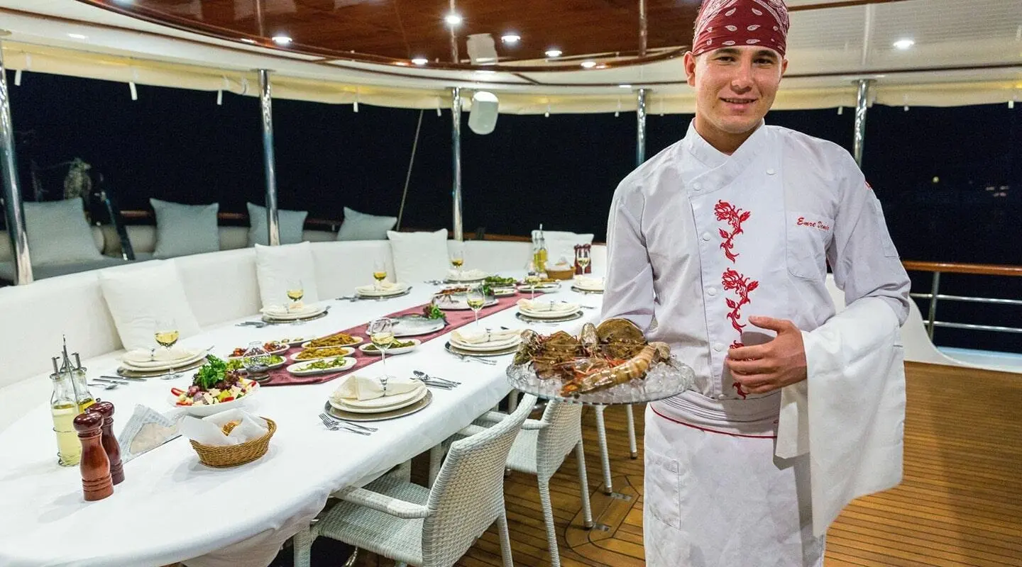 Chef in white uniform standing beside a long dining table set on a yacht deck