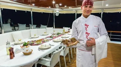 Chef in white uniform standing beside a long dining table set on a yacht deck