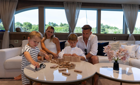 Family playing with wooden blocks on a yacht saloon, white sofas and large windows with green trees outside