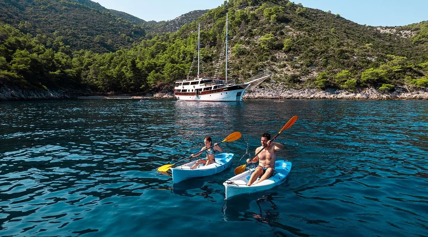 Two people on jet skis near a blue yacht anchored by rugged green cliffs