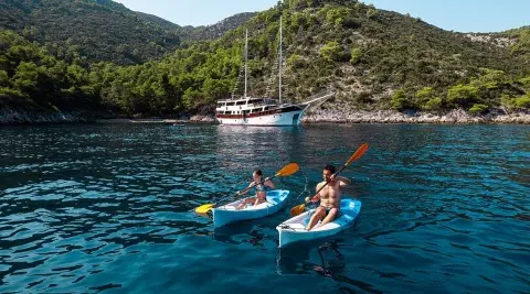 Two people on jet skis near a blue yacht anchored by rugged green cliffs