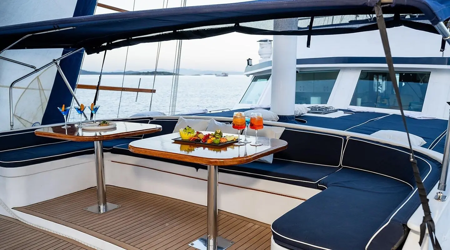 A luxury yacht aft deck with a small wooden table set with drinks and fruit, blue seating, sunset light